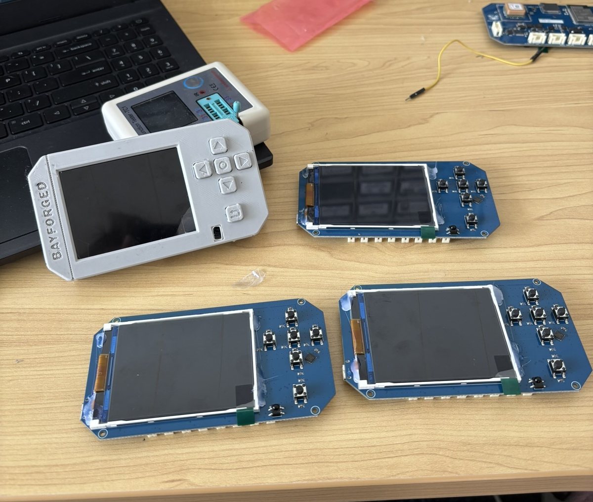 Three bare circuit boards with screens and buttons on a desk, beside one in a white 3D-printed case